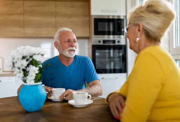 Senior couple sitting at dining table in kitchen, drinking coffee and talking