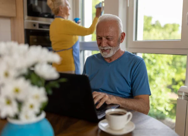 Senior Man Werkt Laptop Aan Eettafel Thuis Terwijl Vrouw Schoonmaken — Stockfoto