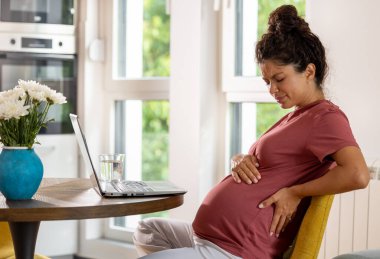Önünde dizüstü bilgisayarla masada oturan hamile bir kadın ve aşırı çalışmanın ardından sırt ağrısı çekiyor.