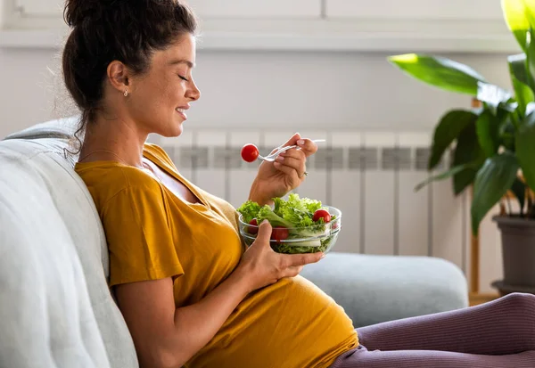 若い女性が自宅でソファでサラダを食べています 産後の健康的な生活 — ストック写真