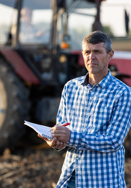 Mature farmer standing in field in front of tractor in autumn and writing notes