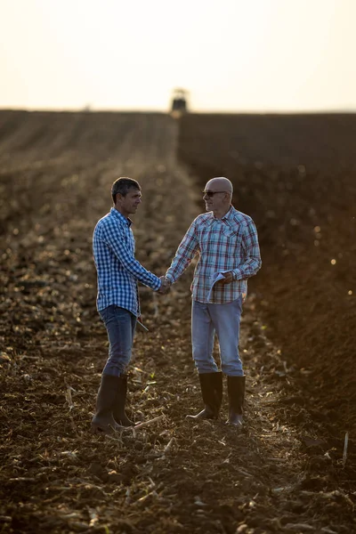 两个高年级的农民在田里握手 而拖拉机则在地里干活 — 图库照片
