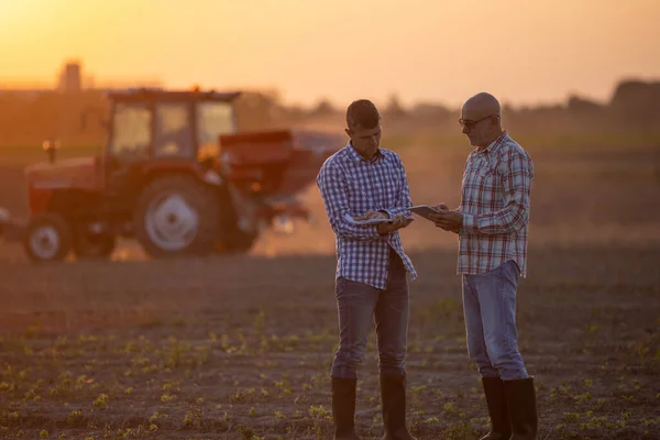 Two senior farmers talking in field in front of tractor at sunset in fall