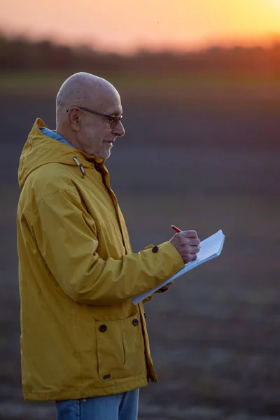 Senior farmer standing in field in dusk and writing notes in late september