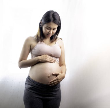 Güzel Asyalı hamile kadın pencerenin yanında karnını gösteriyor. Annem dokunuyor ve elleriyle karnını okşuyor. Annelik, hamilelik, jinekoloji ve sağlık konsepti.