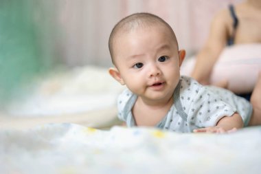 Mutlu aile, tatlı Asyalı yeni doğmuş bebek yatakta bir şeye bakarak oyun oynuyor. Annen yakınlarda sevgiyle ilgilenirken. Masum küçük bir çocuk, hayatının ilk gününde. Anneler Günü konsepti.