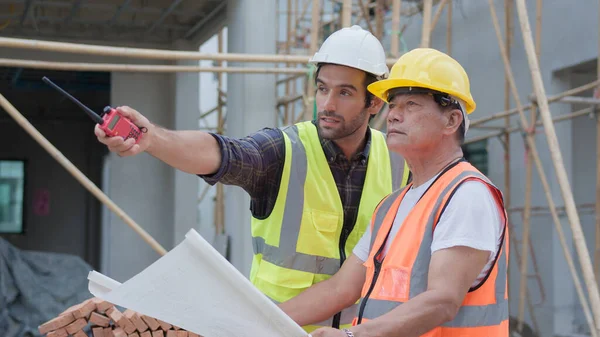 Professional male civil engineer or Architect, inspector with contractor, foreman or worker. While look at blueprint plan inspect discuss meeting building project and pointing on construction site.