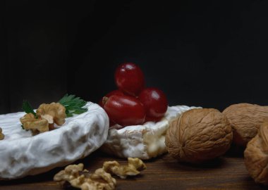 A circle of camembert cheese with white noble mold lies on a wooden board. A piece of cheese with walnuts and grapes.
