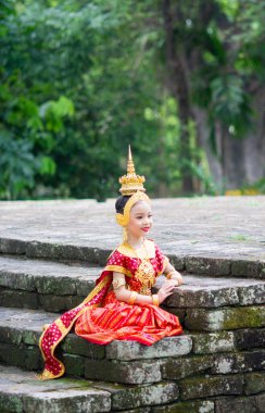 Tipik, geleneksel Tayland elbisesi giyen Asyalı genç kız antika taş bir merdivenin üzerinde oturuyor. Kırmızı ve altın Ulusal kostüm