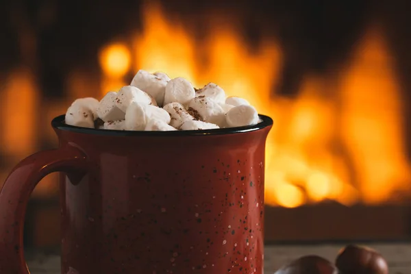 Kakao mit Marshmallows und Schokolade in einem roten Becher auf einem Holztisch neben einem brennenden Kamin — Stockfoto