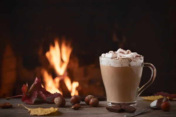 Kakao mit Marshmallows und Schokolade im Glasbecher auf einem Holztisch neben einem brennenden Kamin — Stockfoto