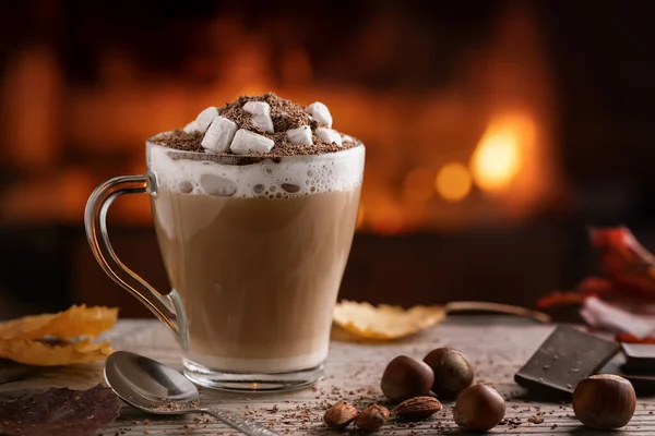 Kakao mit Marshmallows und Schokolade im Glasbecher auf einem Holztisch neben einem brennenden Kamin — Stockfoto