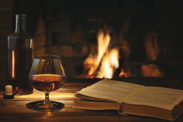 Bicchiere di cognac, una bottiglia e un vecchio libro aperto sul tavolo vicino al camino ardente. Concetto di riposo e relax Immagine Stock