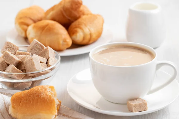 Kaffee mit Milch in einer weißen Tasse, Brötchen und braunem Zucker auf einem weißen Holztisch — Stockfoto
