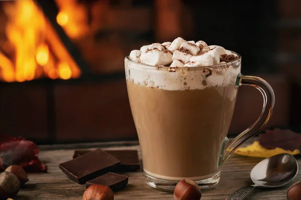 Cacao met marshmallows en chocolade in een glazen mok op een houten tafel bij een open haard — Stockfoto