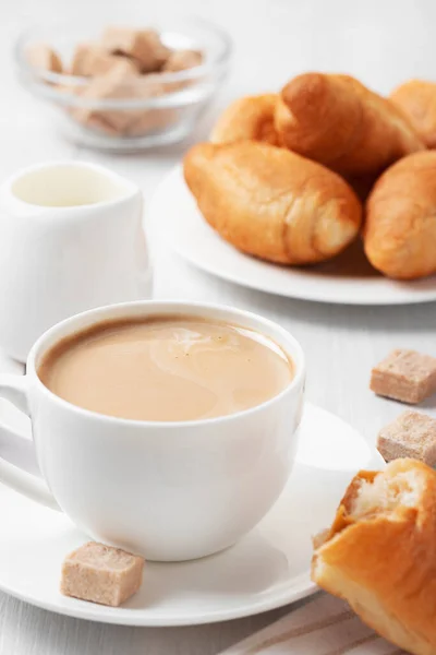 Koffie met melk in een witte beker, broodjes en bruine suiker op een witte houten tafel — Stockfoto