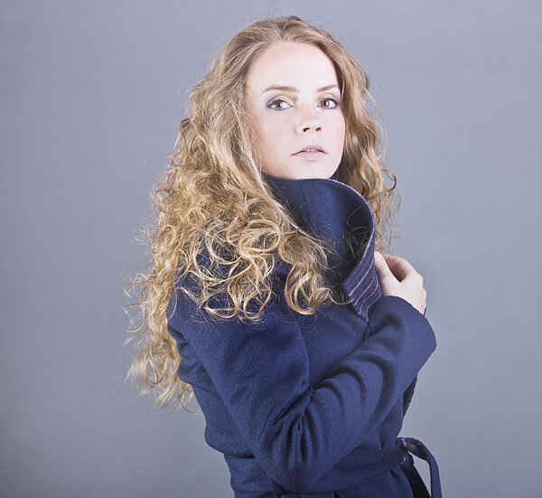 Beautiful curly blonde with a languid sight in the blue coat on gray background