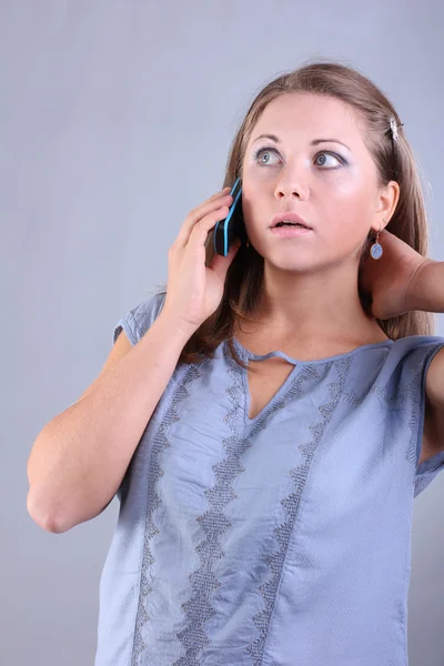 Красивая молодая девушка с голубыми глазами говорит по мобильному телефону на сером фоне — стоковое фото