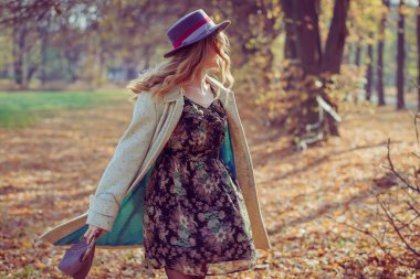 Beyaz önlüklü, ince şapkalı, omuzlarında kırmızı çantalı mutlu kıvırcık kız, Sunn 'daki sonbahar parkında sarı yaprakların ortasında döner.