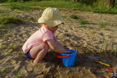 Küçük bebek kumdan arkaplanda suyla dolu mavi plastik bir kovayla oynuyor. Kovadan çıplak ayakla su döken bir çocuk tatilde eğlenceli bir çocukluk dönemi öğreniyor.