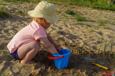 Küçük kız ellerini kumsalda mavi bir plastik kovaya daldırıyor, ellerini kumla yıkıyor mutlu bir çocuklukta güneşli yaz havasındaki yeni keşifler fotoğrafıyla birlikte ellerini yıkıyor.