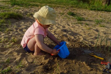 Küçük bebek kumdan arkaplanda suyla dolu mavi plastik bir kovayla oynuyor. Kovadan çıplak ayakla su döken bir çocuk tatilde eğlenceli bir çocukluk dönemi öğreniyor.