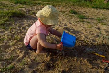 Küçük bebek kumdan arkaplanda suyla dolu mavi plastik bir kovayla oynuyor. Kovadan çıplak ayakla su döken bir çocuk tatilde eğlenceli bir çocukluk dönemi öğreniyor.