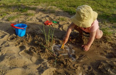 Kumsalda sarı şapkalı küçük bir kız pembe tişörtlü ve mavi şortlu bir kızla eğleniyor. Çok mutlu ve yazın güneşli havayla ilgileniyor.