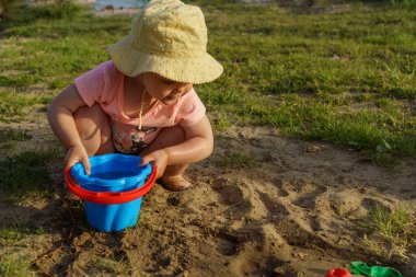 Küçük bebek kumdan arkaplanda suyla dolu mavi plastik bir kovayla oynuyor. Kovadan çıplak ayakla su döken bir çocuk tatilde eğlenceli bir çocukluk dönemi öğreniyor.