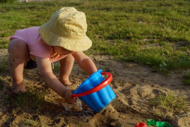 Küçük kız ellerini kumsalda mavi bir plastik kovaya daldırıyor, ellerini kumla yıkıyor mutlu bir çocuklukta güneşli yaz havasındaki yeni keşifler fotoğrafıyla birlikte ellerini yıkıyor.