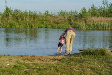 Baba kucağında küçük bir çocuk, yeşil çimlerin etrafındaki göldeki kumdan ayaklarını yıkamaya çalışıyor. Açık deniz, parlak güneş, yaz tatili.