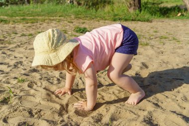 Kumsalda sarı şapkalı küçük bir kız pembe tişörtlü ve mavi şortlu bir kızla eğleniyor. Çok mutlu ve yazın güneşli havayla ilgileniyor.