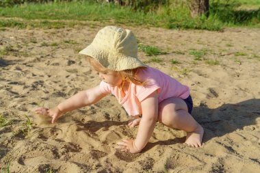 Kumsalda sarı şapkalı küçük bir kız pembe tişörtlü ve mavi şortlu bir kızla eğleniyor. Çok mutlu ve yazın güneşli havayla ilgileniyor.