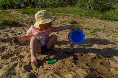 Küçük bebek kumdan arkaplanda suyla dolu mavi plastik bir kovayla oynuyor. Kovadan çıplak ayakla su döken bir çocuk tatilde eğlenceli bir çocukluk dönemi öğreniyor.
