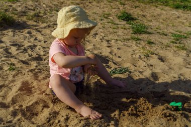 Kumsalda sarı şapkalı küçük bir kız pembe tişörtlü ve mavi şortlu bir kızla eğleniyor. Çok mutlu ve yazın güneşli havayla ilgileniyor.