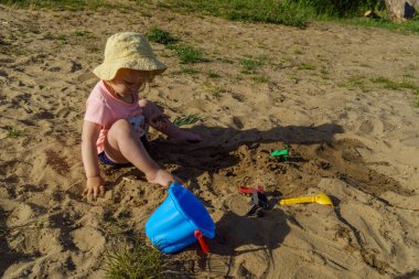 Kumsalda sarı şapkalı küçük bir kız pembe tişörtlü ve mavi şortlu bir kızla eğleniyor. Çok mutlu ve yazın güneşli havayla ilgileniyor.