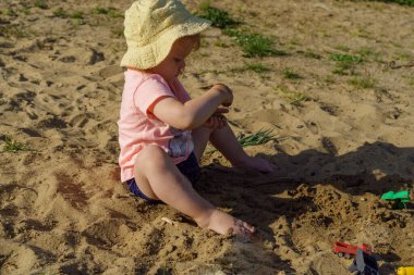 Kumsalda sarı şapkalı küçük bir kız pembe tişörtlü ve mavi şortlu bir kızla eğleniyor. Çok mutlu ve yazın güneşli havayla ilgileniyor.