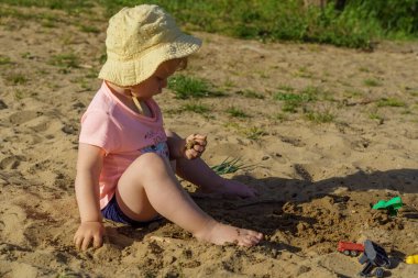 Kumsalda sarı şapkalı küçük bir kız pembe tişörtlü ve mavi şortlu bir kızla eğleniyor. Çok mutlu ve yazın güneşli havayla ilgileniyor.