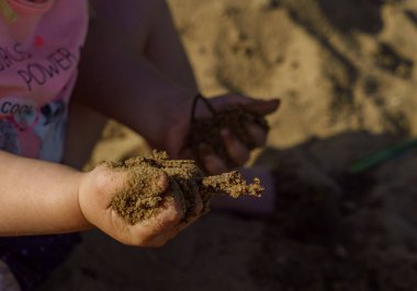 Kumsalda sarı şapkalı küçük bir kız pembe tişörtlü ve mavi şortlu bir kızla eğleniyor. Çok mutlu ve yazın güneşli havayla ilgileniyor.