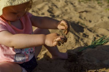 Kumsalda sarı şapkalı küçük bir kız pembe tişörtlü ve mavi şortlu bir kızla eğleniyor. Çok mutlu ve yazın güneşli havayla ilgileniyor.