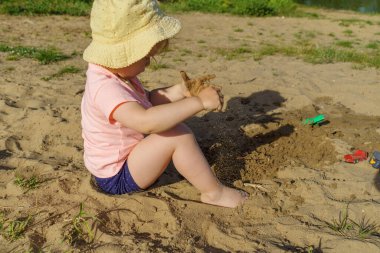 Kumsalda sarı şapkalı küçük bir kız pembe tişörtlü ve mavi şortlu bir kızla eğleniyor. Çok mutlu ve yazın güneşli havayla ilgileniyor.