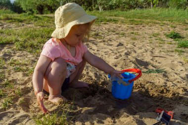 Küçük bebek kumdan arkaplanda suyla dolu mavi plastik bir kovayla oynuyor. Kovadan çıplak ayakla su döken bir çocuk tatilde eğlenceli bir çocukluk dönemi öğreniyor.