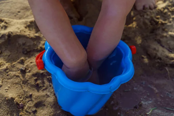 little girl dips her hands in a blue plastic bucket of water on the beach with sand, washes her hands in a bucket happy childhood with new discoveries photo in sunny summer weather