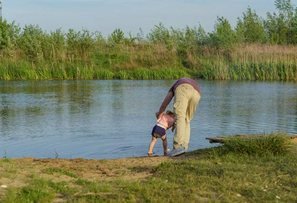 Baba kucağında küçük bir çocuk, yeşil çimlerin etrafındaki göldeki kumdan ayaklarını yıkamaya çalışıyor. Açık deniz, parlak güneş, yaz tatili.