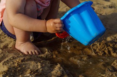 Küçük bebek kumdan arkaplanda suyla dolu mavi plastik bir kovayla oynuyor. Kovadan çıplak ayakla su döken bir çocuk tatilde eğlenceli bir çocukluk dönemi öğreniyor.