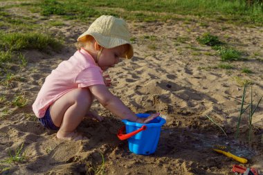 Küçük kız ellerini kumsalda mavi bir plastik kovaya daldırıyor, ellerini kumla yıkıyor mutlu bir çocuklukta güneşli yaz havasındaki yeni keşifler fotoğrafıyla birlikte ellerini yıkıyor.
