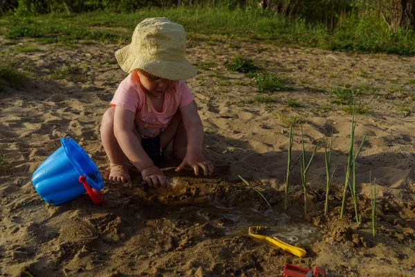 little girl on the beach in a yellow hat having fun playing with sand girl wearing a pink t-shirt and blue shorts girl very happy and interested in sunny warm weather in summer on vacation