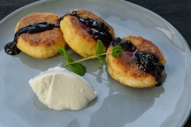 Kızarmış, çıtır çıtır peynirli kekler, kiraz reçeli ile doldurulmuş, gri bir seramik tabakta ekşi kremayla servis edilmiş ve bir tutam kokulu nane ile süslenmiş.