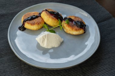 Kızarmış, çıtır çıtır peynirli kekler, kiraz reçeli ile doldurulmuş, gri bir seramik tabakta ekşi kremayla servis edilmiş ve bir tutam kokulu nane ile süslenmiş.