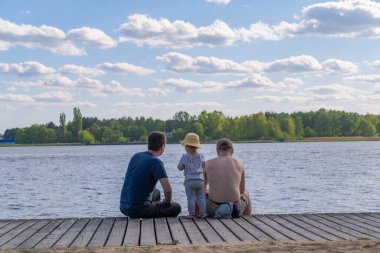  a couple men with a child, sit on the pier facing the water and look into the distance at the water, at the sky with white clouds, the family enjoys rest, the child in a hat, a photo from the back
